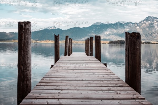 A wooden footbridge leads into a tranquil lake, surrounded by mountains under an open sky
