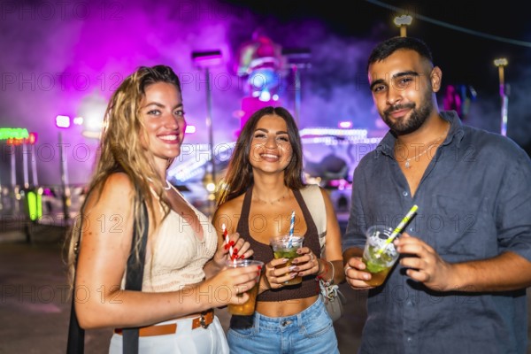 Three friends are holding cocktails while enjoying the vibrant nightlife at an amusement park, illuminated by colorful lights and attractions