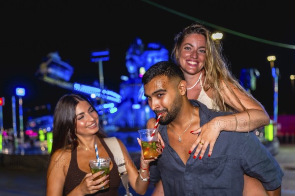 Three cheerful friends enjoying refreshing cocktails at an amusement park, illuminated by vibrant lights, creating a joyful atmosphere
