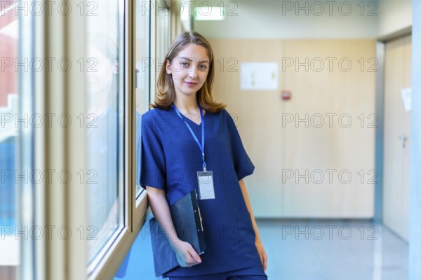 Confident young doctor wearing blue scrubs holding a clipboard while standing in a modern hospital corridor