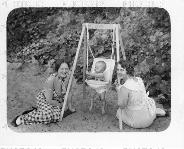 Women with baby on swing, 1930s, Two woman kneeling smiling in front of a baby in a swing, sitting on the beach, Historical photo