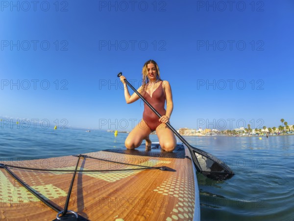 Young woman kneeling on a paddle board, holding a paddle, enjoying a summer watersport activity on a sunny day with clear blue sky