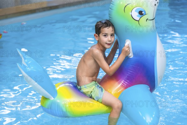 Smiling boy riding a colorful inflatable sea lion toy while splashing in the swimming pool during a joyful summer vacation day