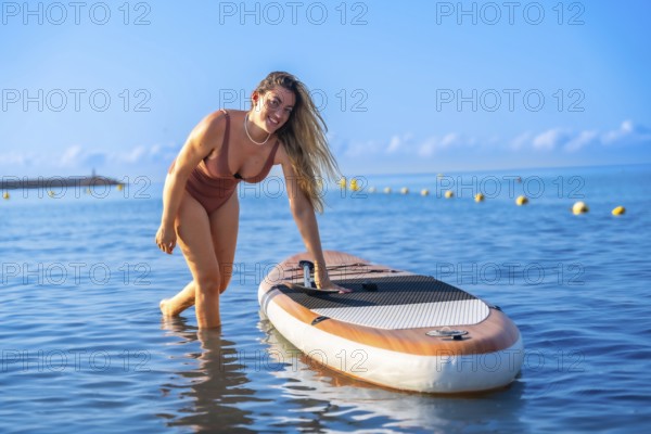 Woman engaging in paddle surfing on a calm summer day, standing beside her paddleboard in clear blue water, showcasing outdoor leisure activities