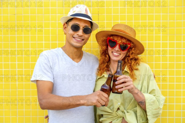 Happy couple toasting with beer bottles while enjoying a summer day against a vibrant yellow tiled wall