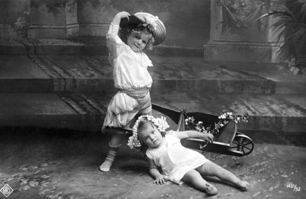 Child falls out of wheelbarrow, 1910, Two children playing in the garden, one with a wreath of flowers lies in a wheelbarrow, Historical photo