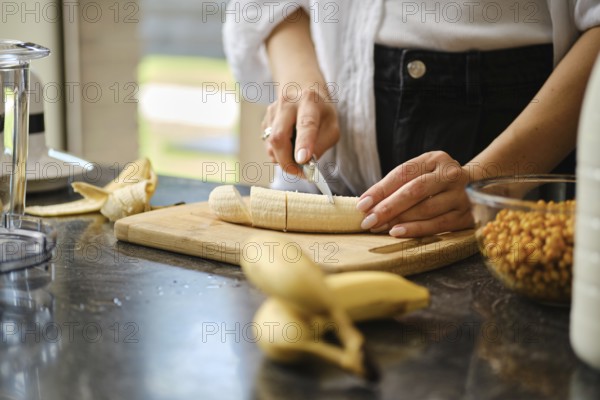 In a warm kitchen, a person slices ripe bananas on a wooden cutting board. Fresh sea buckthorn berries and other ingredients are visible, showcasing the preparation for nutritious smoothies
