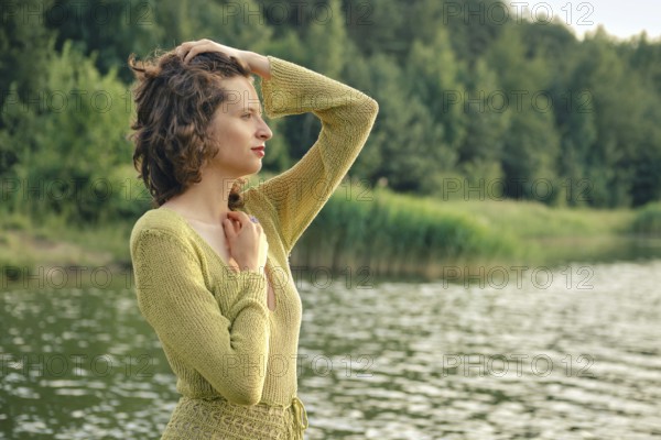 On a hot summer day, a woman stands by the tranquil lake, her hand resting on her head while she gazes thoughtfully at the shimmering water. Lush greenery surrounds the scene, enhancing the beauty