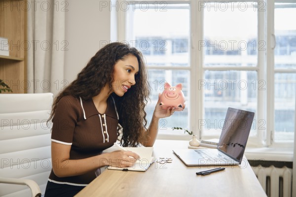 A young woman is actively budgeting at her desk, holding a pink piggy bank in one hand while writing notes in a notebook with her other hand