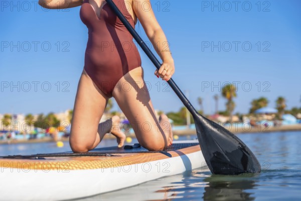 Woman kneeling on a paddleboard, holding a paddle on a calm summer day. Clear blue sky and distant beach in the background