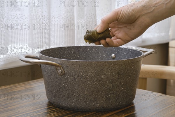A person prepares traditional dolma by carefully placing a stuffed grape leaf into a pot on a wooden table, surrounded by soft natural light from a window. The atmosphere exudes warmth and comfort