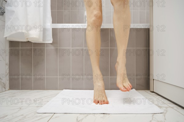 A person is standing on a fluffy white towel in a bathroom, drying wet feet after taking a shower. Light reflects off smooth gray tiles, creating a serene atmosphere filled with cleanliness