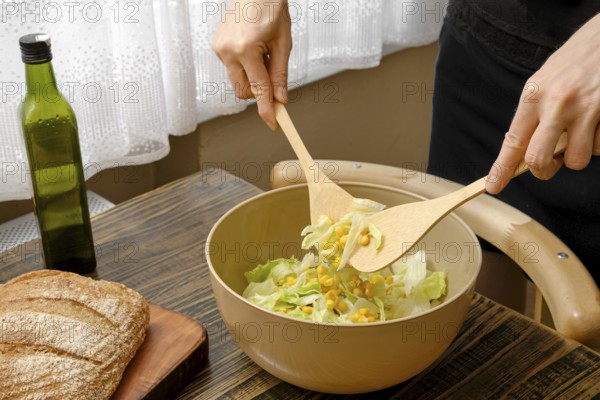 A person stirs a vibrant salad with wooden spoons in a warm and inviting kitchen. Fresh lettuce and corn are being combined in a large bowl, while a bottle of olive oil and some bread sit nearby