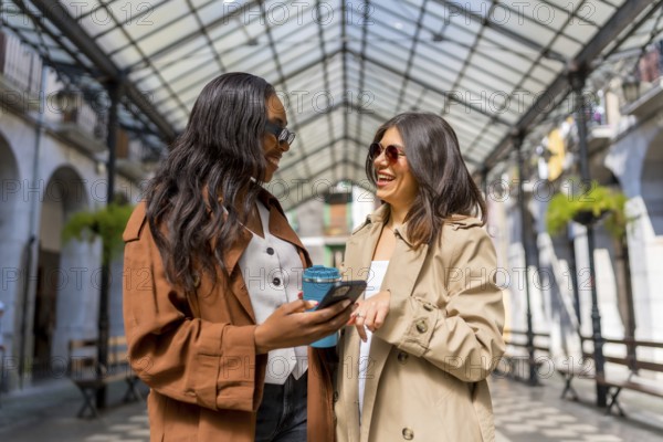 Two cheerful businesswomen are enjoying a moment together, using a smartphone and holding a reusable coffee cup in a covered gallery