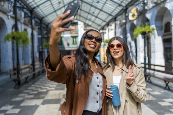 Two female friends taking a selfie in a covered walkway in a city center, one is holding a reusable cup
