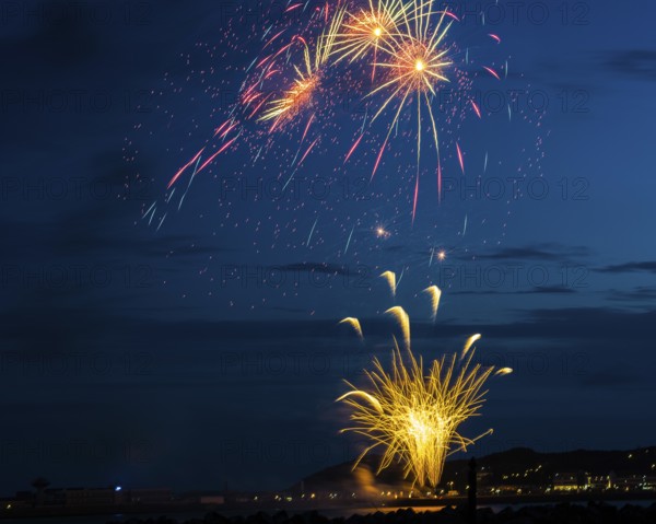 A brilliantly radiant, atmospheric firework display for the Heligoland island festival in red and gold over the night-time lowlands, inland harbour, jetty and lobster shacks, with the reflecting waters of the North Sea in front, golden-coloured fire fountain and three whirling stars, Night sky at the island festival in summer, symbolic image also for New Year's Eve, New Year's Eve fireworks, bright, colourful, many colourful traces of light, sparks, lively glitter, night, Heligoland Island, Schleswig-Holstein, Germany