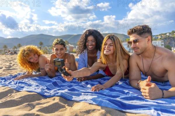 Five young friends lying on a striped towel on a sandy beach, taking a selfie with an action camera