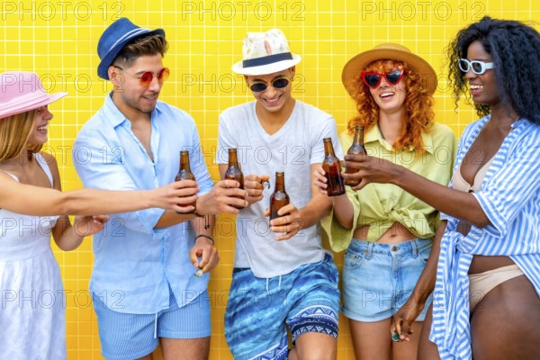 Group of cheerful young friends toasting with beer bottles while standing together against a vibrant yellow tiled wall, enjoying their summer vacation