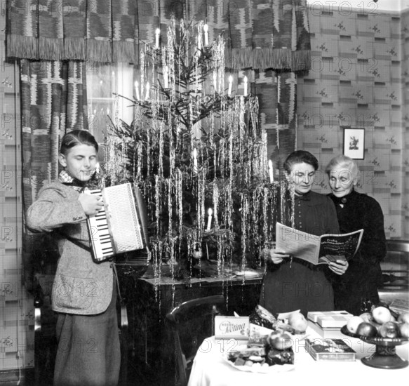 Young-mother-grandmother-Christmas, 1940s, Three generations celebrate Christmas next to a festive tree with musical accompaniment, Historical photo