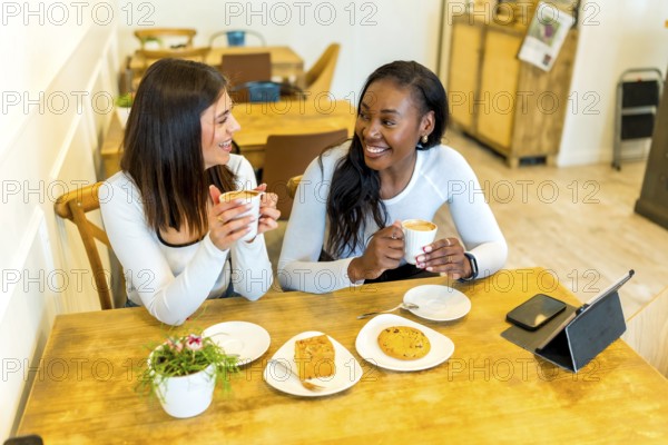 Two female friends enjoying coffee and snacks while chatting and laughing together at a cozy cafe table, sharing a delightful moment