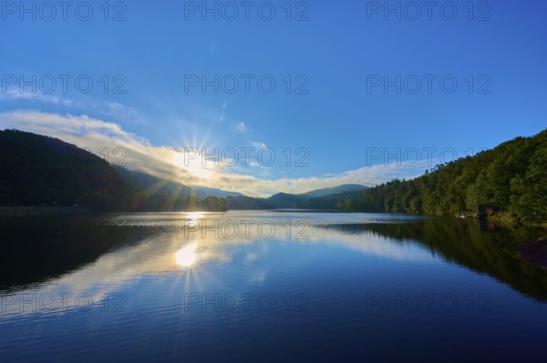 The bright morning sun illuminates the sky and is reflected on a calm lake surrounded by rolling hills, summer, Rursee, Obersee, Rurberg, Simmerath, Eifel National Park, Eifel, North Rhine-Westphalia, Germany