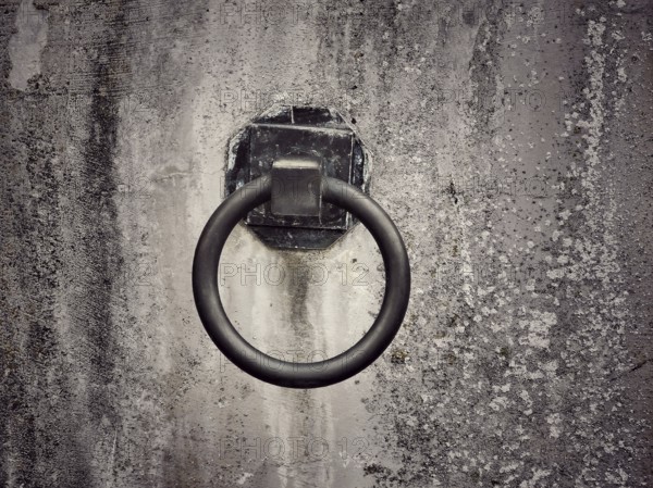 A weathered wall features a rusty metal ring affixed to it, suggesting a historical purpose. The texture and color highlight the passage of time and use in an urban setting