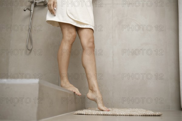 A woman is stepping carefully onto a soft bath mat from a tiled shower area in a sleek and contemporary bathroom setting. The light tones create a calming atmosphere