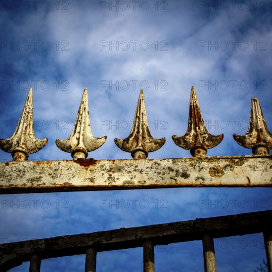 Close-up view of an ornamental iron fence featuring pointed spikes, showcasing rust and weathering against a dramatic cloudy sky in a garden setting. France