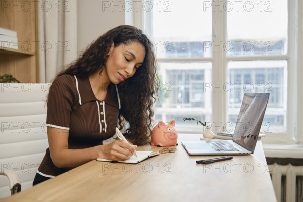 A woman sits at a wooden desk, writing in a notebook and planning her finances in a bright home office. A laptop and a pink piggy bank are visible, suggesting a focus on saving and organization
