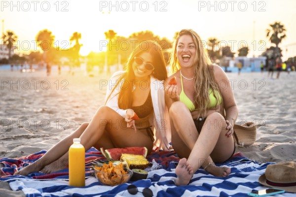 Two female friends enjoying a picnic with fresh fruit and juice on a beach towel at sunset, creating a joyful summer scene