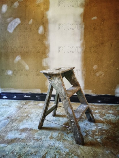 A worn wooden ladder rests on an uneven floor in a room with a freshly painted wall. The surroundings indicate ongoing home improvement work, suggesting preparation for completion
