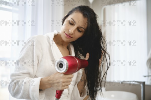 A woman with long dark hair is drying her hair using a red hairdryer while wearing a white robe. She stands in a well-lit bathroom, focused on getting her hair dry and styled