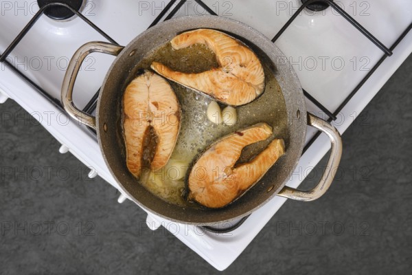 Salmon fillets are being cooked in a pan with garlic on a stovetop. The kitchen is well-lit and the fillets show a golden color, indicating they are almost ready to be served