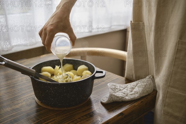 A hand pours warm milk onto a pot filled with boiled potatoes on a wooden table. The process of making mashed potato at home