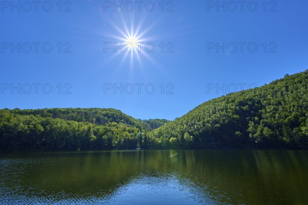 Bright sun over a calm lake with green hills and blue sky, summer, Rursee, Obersee, Simmerath, Eifel National Park, North Rhine-Westphalia, Germany