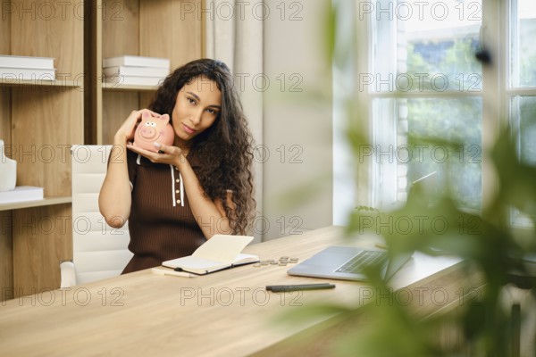 In a contemporary office space, a woman with curly hair sits at a wooden desk, smiling as she holds a piggy bank. A notebook and laptop are visible, indicating she is planning her finances