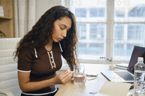 A woman with curly hair sits at a desk in a contemporary office, holding a pill and a glass of water