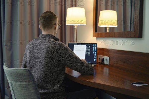 A person sits at a wooden desk in a hotel room, focused on his laptop screen. Soft lamp light creates a warm ambiance as he reviews documents late in the evening