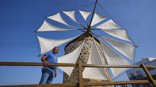 A man leaning against a wooden fence in front of a windmill with white sails and a building in the background, Windmill, Antimacheia, Kos, Dodecanese, Greek Islands, Greece