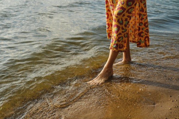 Unrecognizable woman walks barefoot along a sandy beach as waves gently lap at her feet. She wears a vibrant dress with a bold pattern, and the sun sets, casting warm hues on the water
