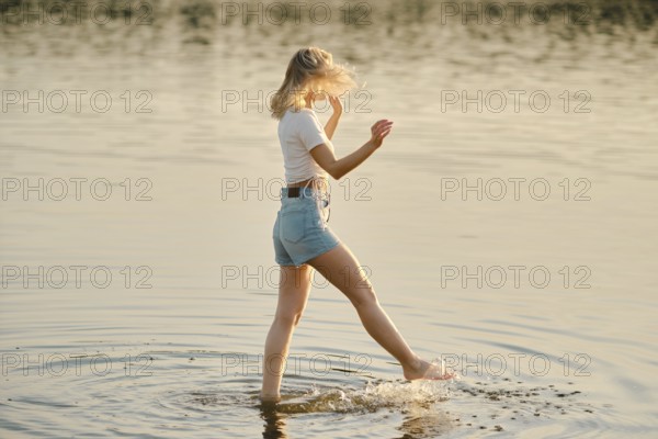 A woman walks through shallow water, splashing lightly as golden sunlight reflects off the surface. The calm setting creates a peaceful summer atmosphere during the evening hours