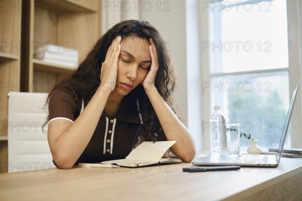 Tired young woman sits at her desk, visibly stressed and holding her head in her hands. The workspace features a notebook, a laptop, and a water bottle, with natural light coming from a window