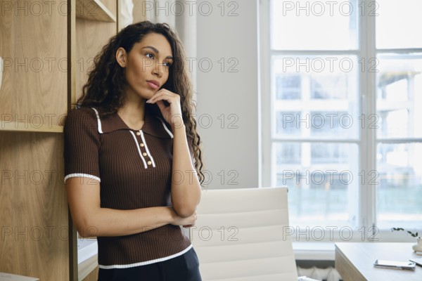 In a modern office, a woman with curly hair stands pensively against a wooden shelving unit