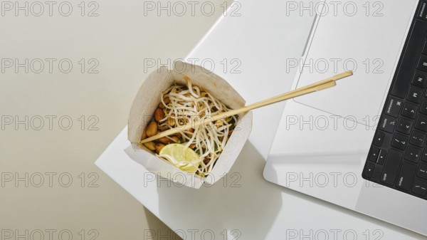 A cardboard bowl of noodles with vegetables and lime sits next to a laptop on a white table. The setting suggests multitasking, combining dining and work in a contemporary environment