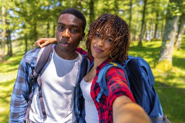 Two young hikers with backpacks smiling while taking a selfie in a sunlit forest, capturing the joy of their outdoor adventure together