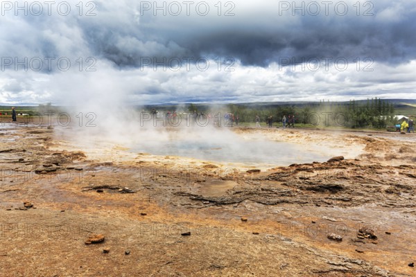 Steaming Strokkur, tourists waiting for eruption, high temperature area Haukadalur, tourist attraction at the Golden Circle, dark rain clouds, Sudurland, Iceland