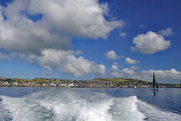 View of the sea with wave foam and boats in front of a town under a blue sky, Stromness, Mainland, Orkney Islands, Scotland, United Kingdom