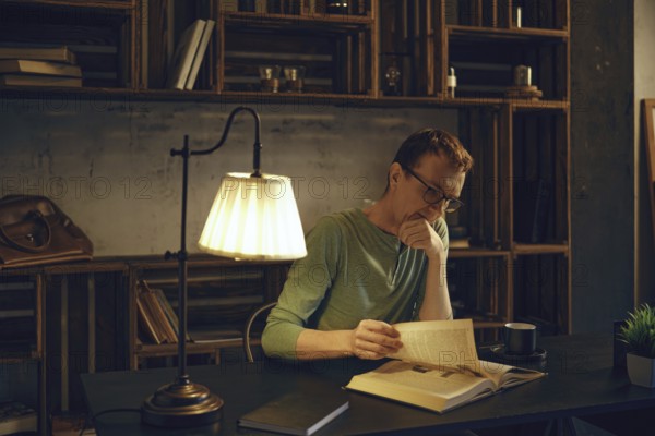 A middle-aged man in glasses sits at a dark wooden desk, deeply engaged in a book