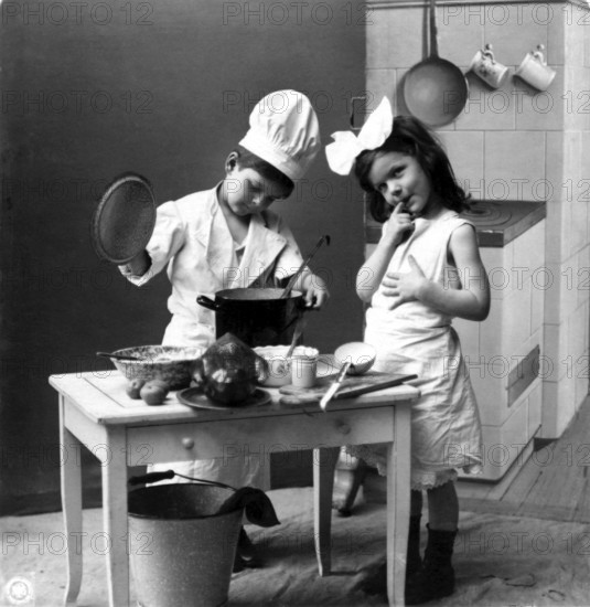 Two children cooking, 1920s, Two children playing in a kitchen, one in a cook's costume, in a black and white photograph, Historical photograph