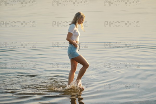 A young woman wearing a white t-shirt and denim shorts walks slowly through the shallow waters of a lake. The sun sets in the background, creating a peaceful atmosphere with gentle ripples